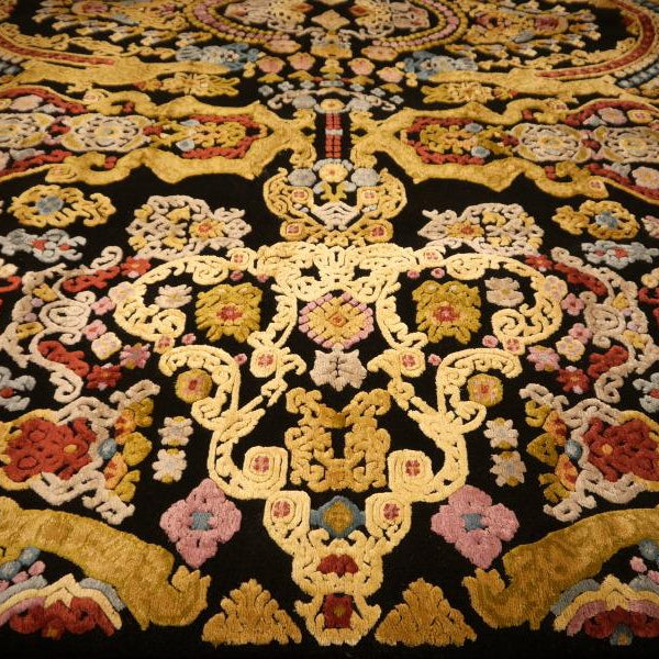 A close-up of an ornate traditional rug with a black background and intricate multicolor floral and scroll patterns in gold, beige, red, pink, and blue.