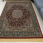 A traditional Persian-style area rug with a dark red central field, intricate floral motifs, and a red border, shown on a white floor in front of a gray sectional sofa.
