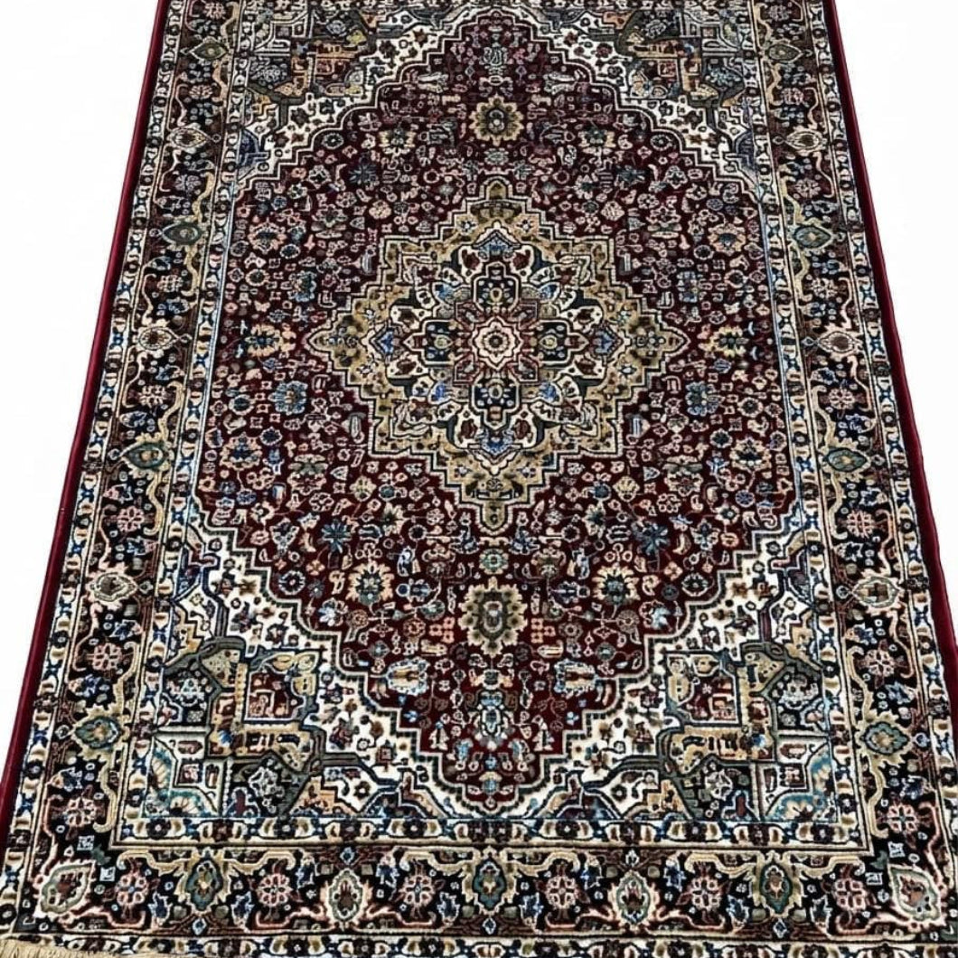 A traditional oriental area rug with an intricate floral and geometric pattern, featuring a dark red central field, beige and blue borders, and cream-colored fringe on the top and bottom edges.