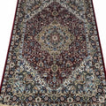 A traditional oriental area rug with an intricate floral and geometric pattern, featuring a dark red central field, beige and blue borders, and cream-colored fringe on the top and bottom edges.