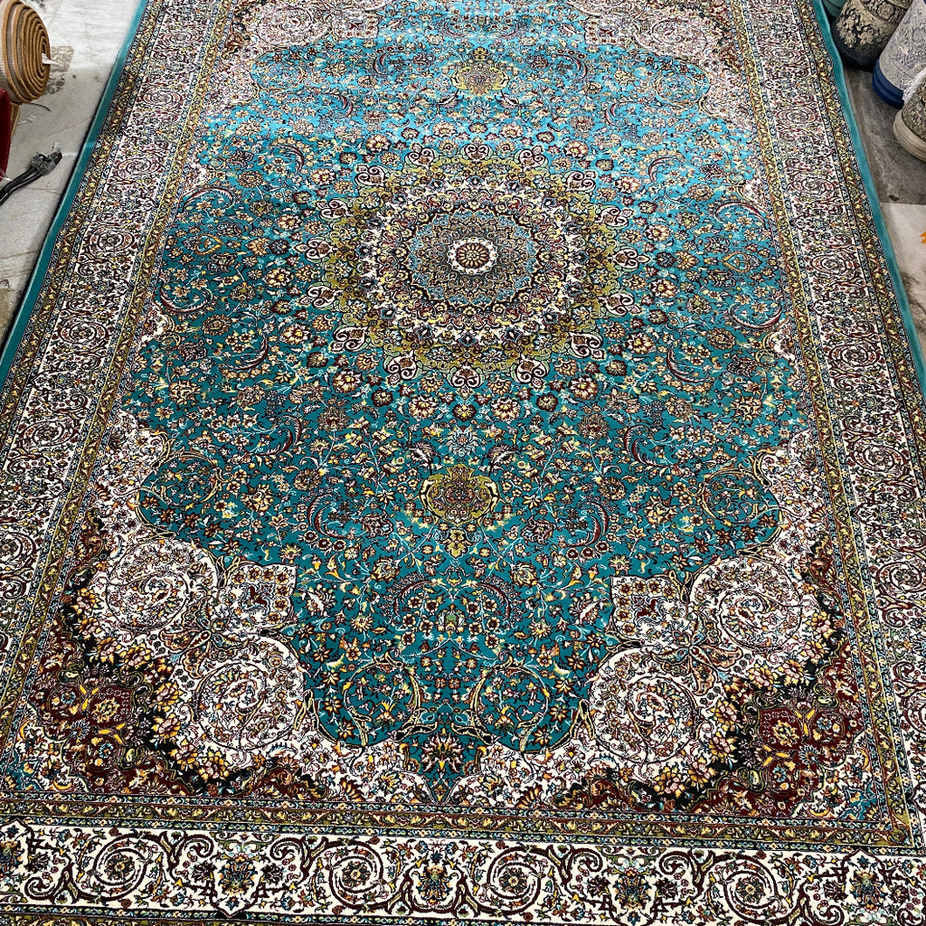 A traditional Persian-style area rug with a vibrant turquoise blue center, intricate floral patterns, and beige fringe, photographed flat on a workshop floor.
