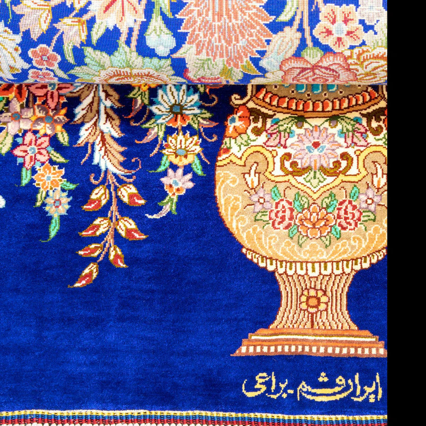 A close-up of a richly patterned rug featuring a large ornate golden vase with floral motifs on a deep blue background, with additional floral vine patterns and Persian script near the bottom.