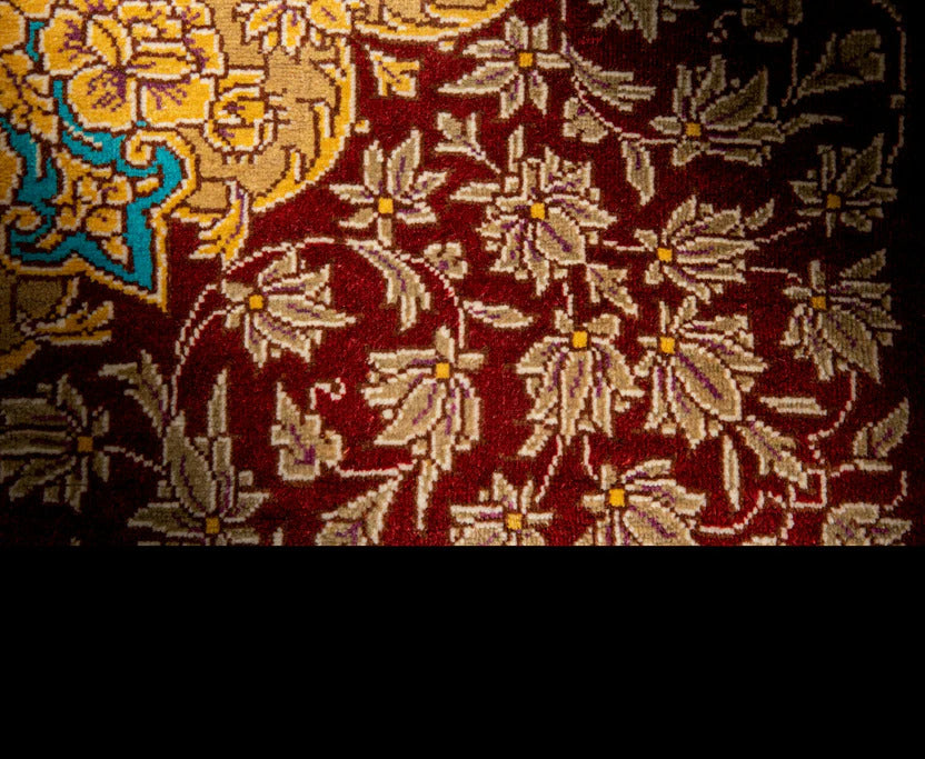 A close-up of a patterned textile or rug showing a deep red field with beige floral motifs and a small blue and yellow decorative section in the upper left.