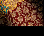 A close-up of a patterned textile or rug showing a deep red field with beige floral motifs and a small blue and yellow decorative section in the upper left.