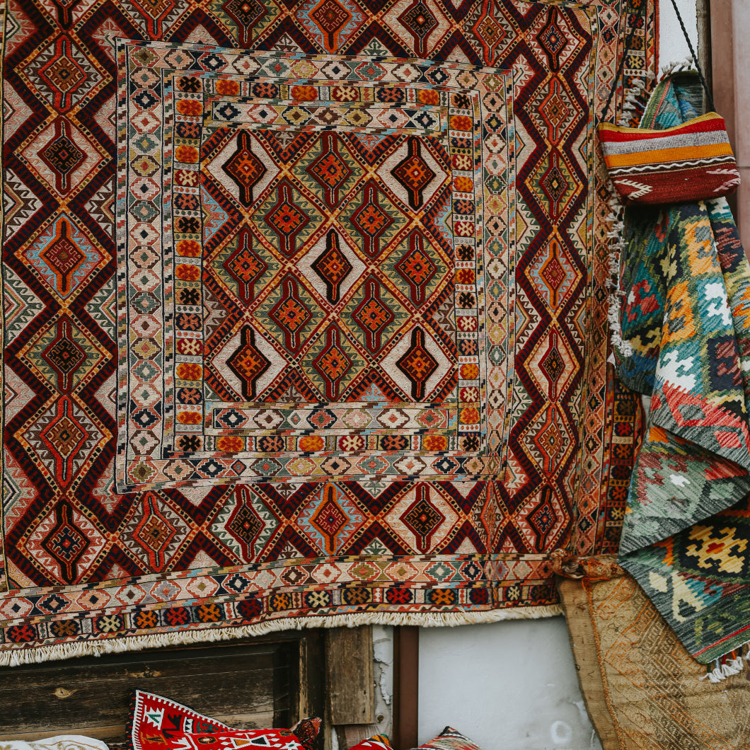 A large traditional geometric area rug hangs on a display stand, featuring a complex pattern of red, orange, beige, and black. Other colorful woven textiles are partially visible on the right and bottom.