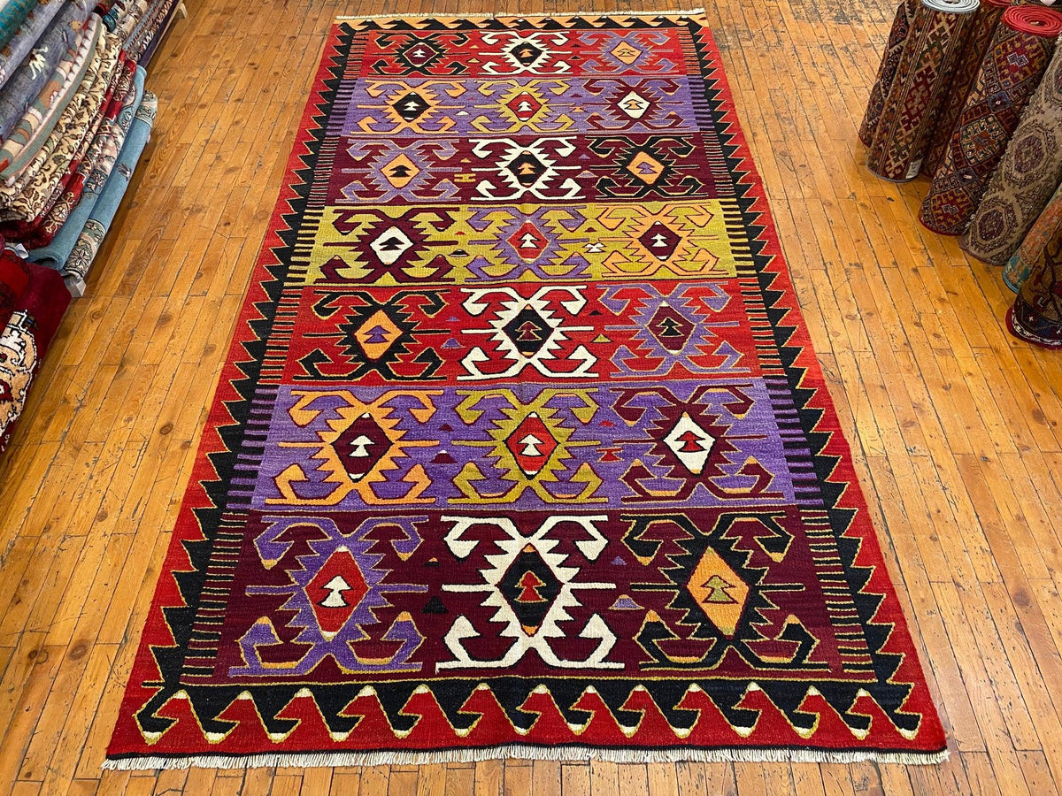 A rectangular woven area rug with a bold geometric tribal pattern in red, purple, yellow, black, and white, displayed on a light wood floor.